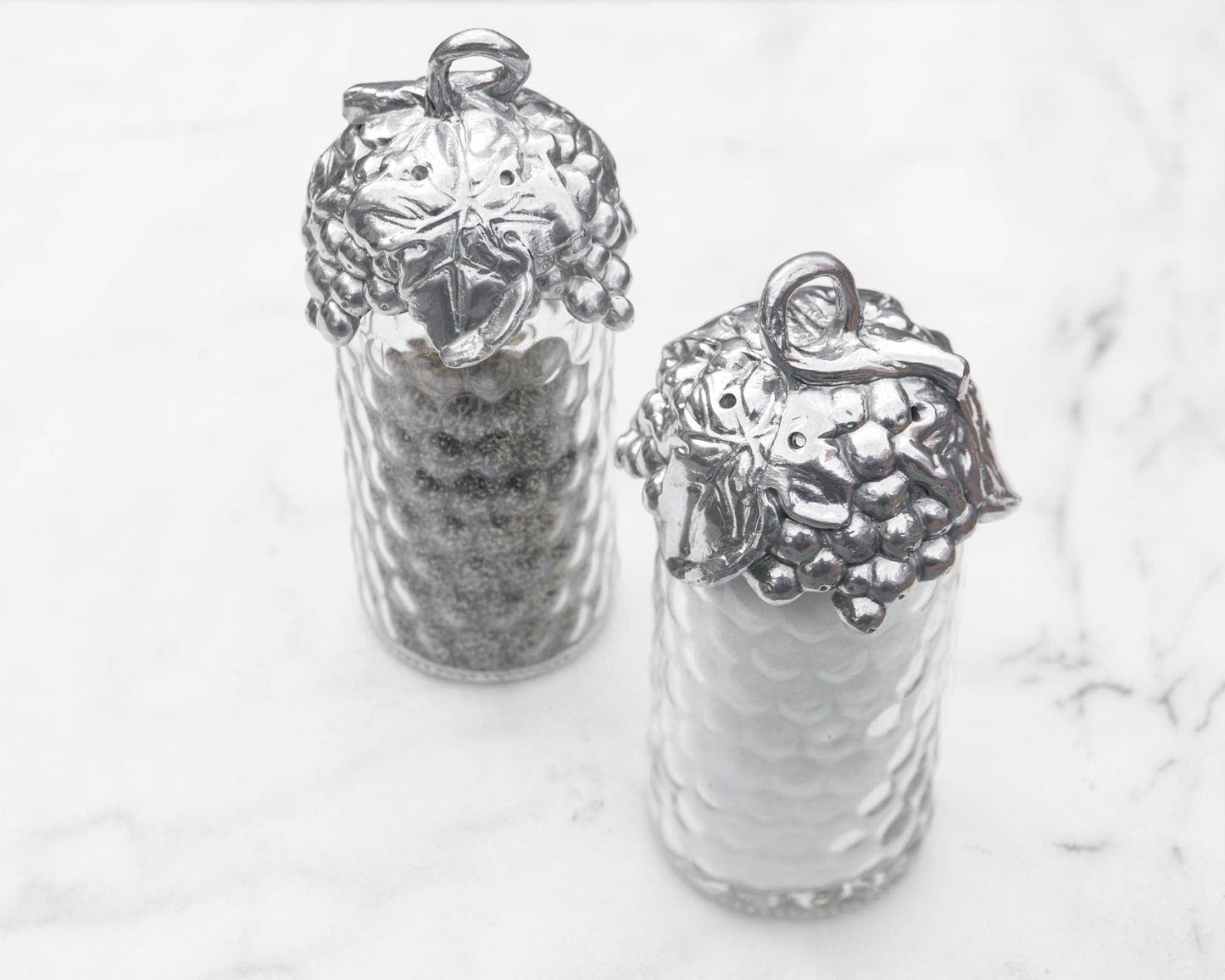 "Grape Salt and Pepper Set"Add a touch of sophistication to your table with this Grape Salt and Pepper Set from Arthur Court. The top of these dispensers features a looping vine adorned with leaves and grapes, creating a unique and elegant design. Handcrafted from aluminum, these pieces will elevate any setting with their simple yet luxurious appearance. Measuring at a diameter of 2.0" and a height of 5.75", these easily refillable dispensers can be dusted with a damp cloth and will not tarnish over time"